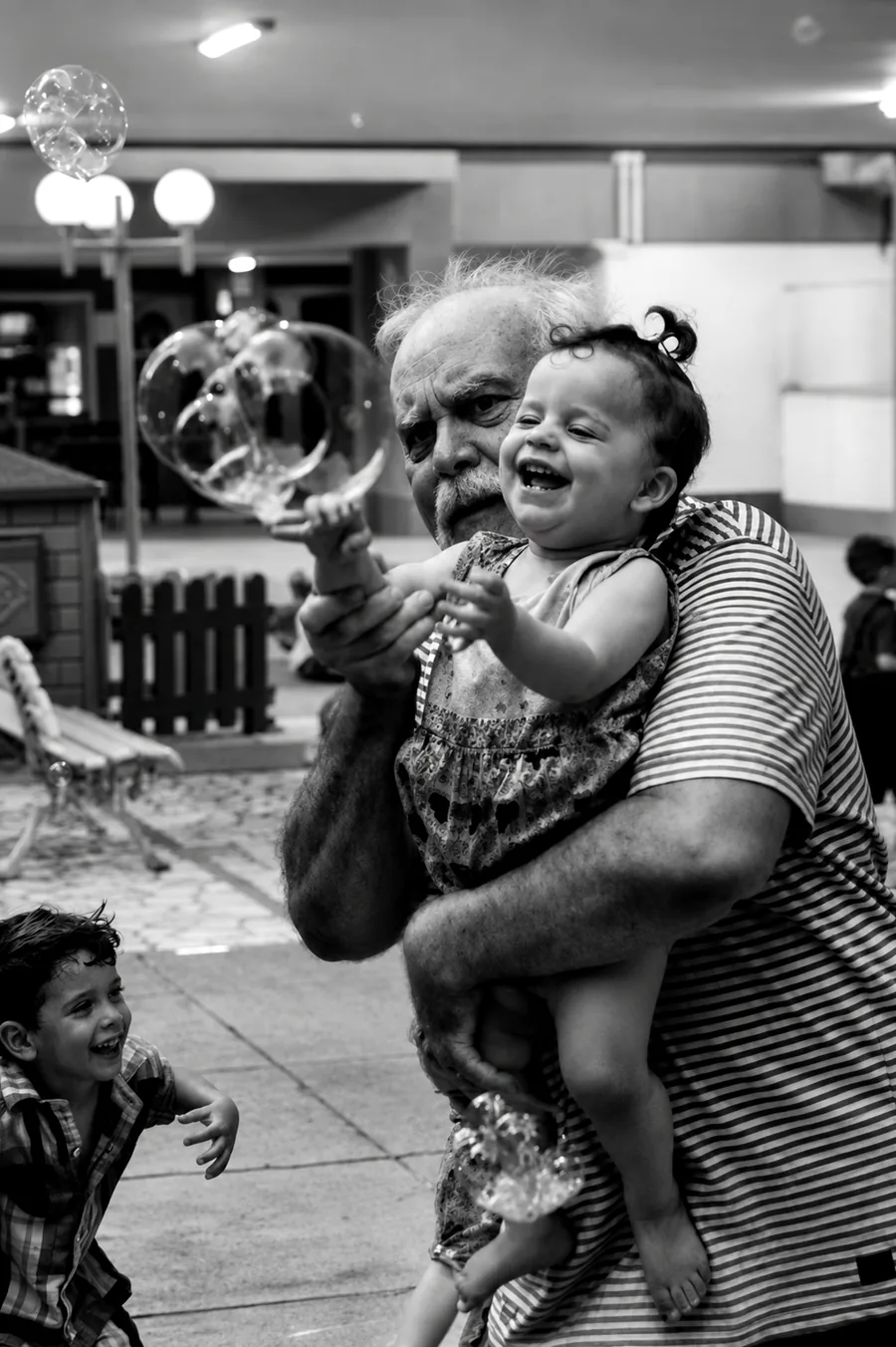 Fotografia de festa infantil no Rio de Janeiro em preto e branco: avô com a neta no colo rindo entre bolhas de sabão, fotógrafo Pirralhitus RJ